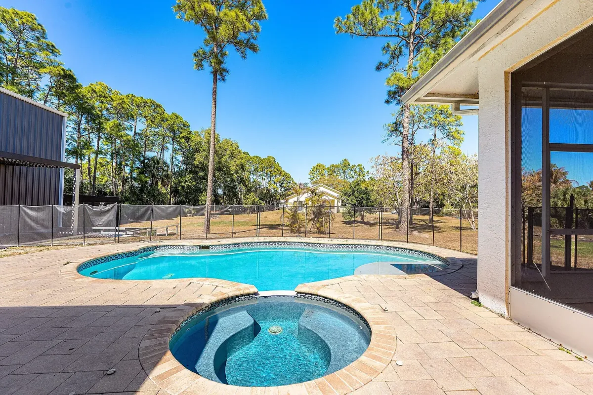 In-ground pool and spa on a paver deck, with the steel building beyond the tree line